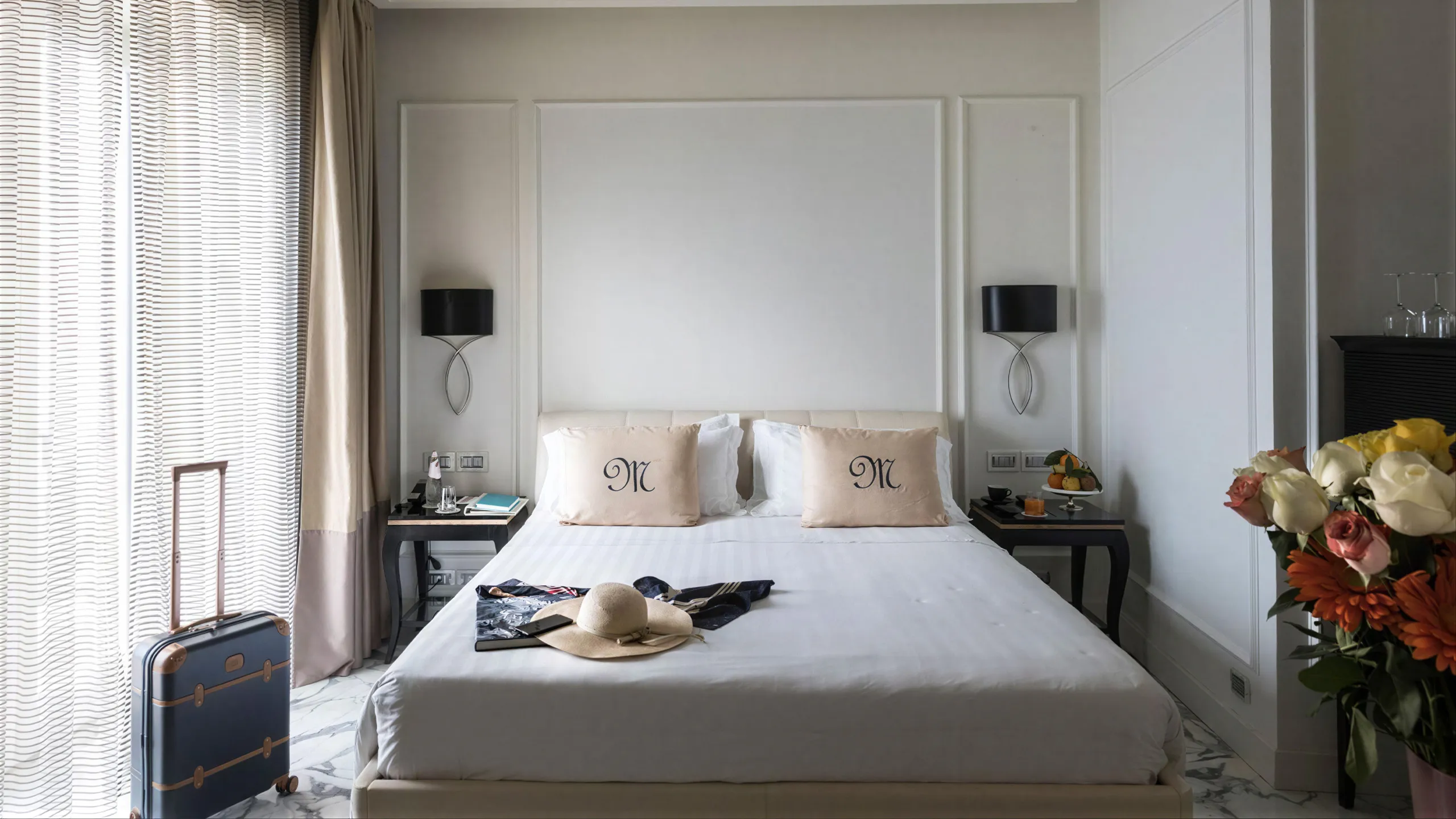 Camera Deluxe vista mare: letto matrimoniale con cuscini monogramma, lampade nere e valigia trolley in primo piano