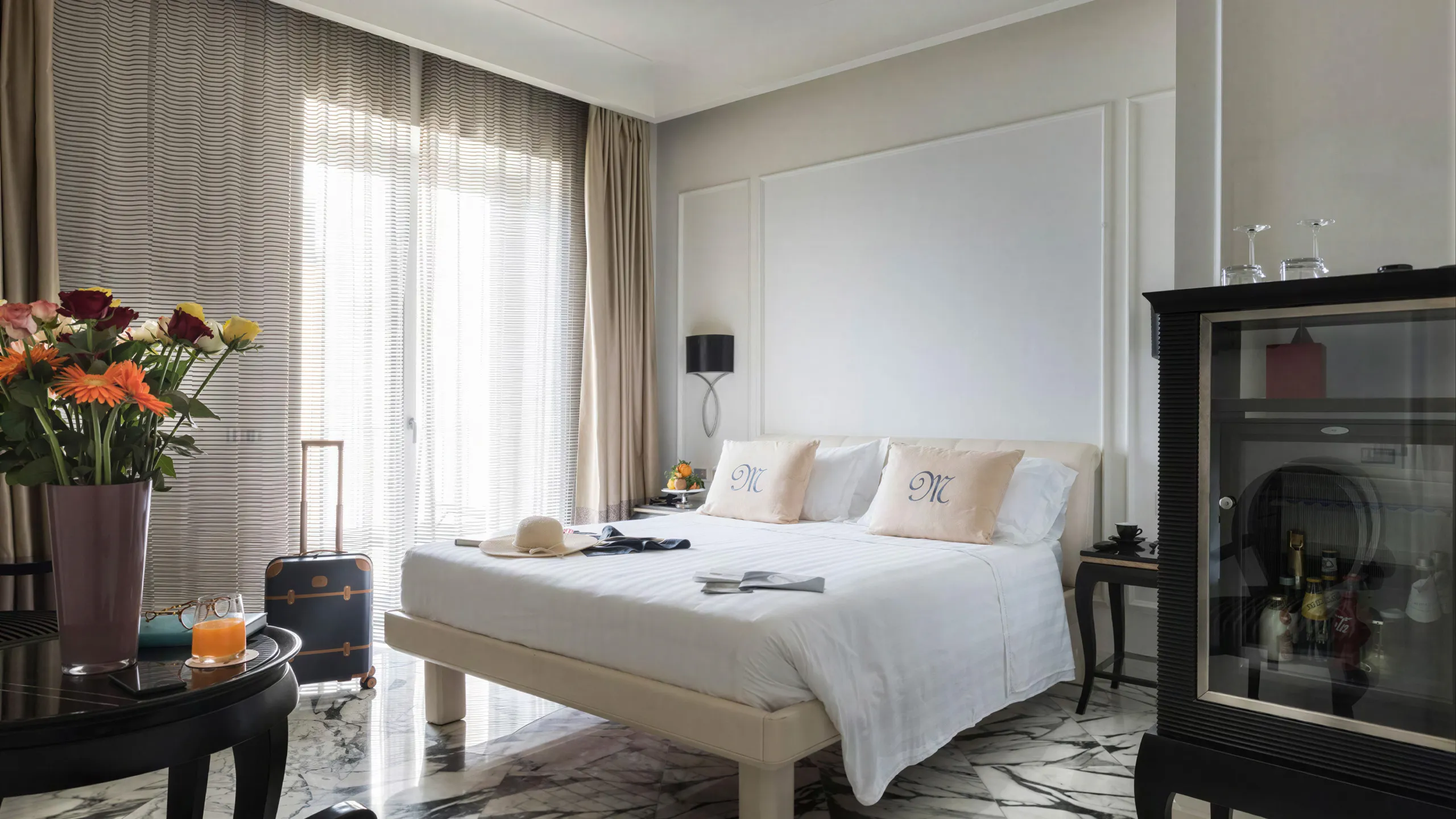 Sea view Deluxe Room: bed with monogram pillows, colourful flowers, minibar and black and white marble floor