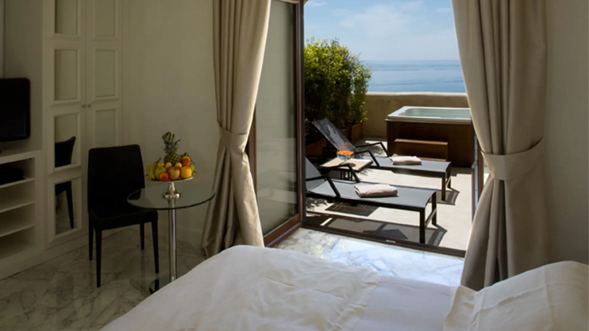 Deluxe Pool Room with terrace: interior with fresh fruit, open door onto sun loungers, jacuzzi and Ionian Sea view