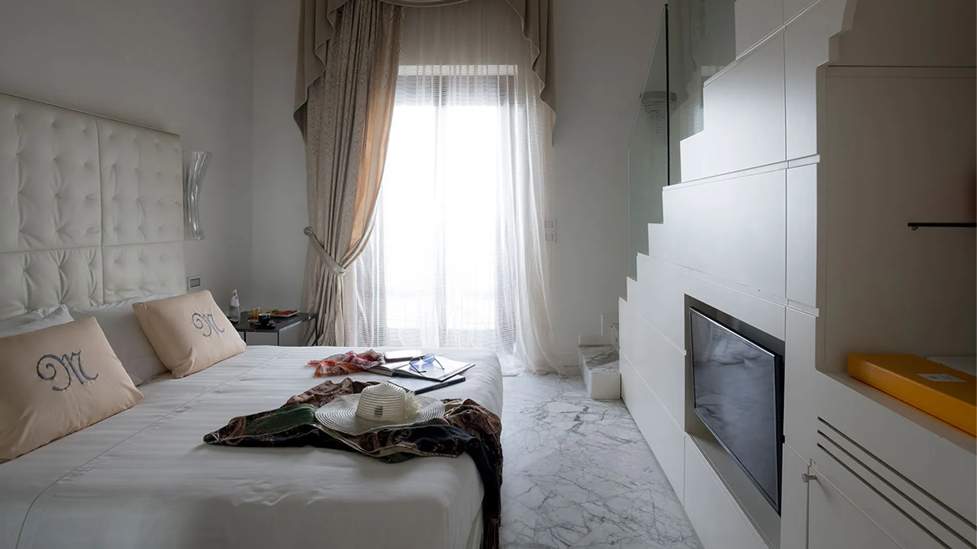 Two-level Junior Suite: bed with capitonné headboard and monogram pillows, white marble floor and internal staircase