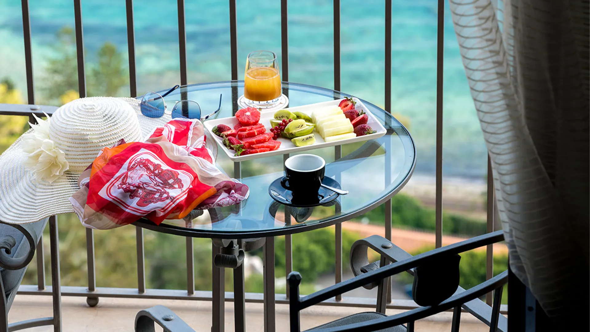 Junior Suite balcony: glass table with fresh fruit, coffee and orange juice, summer hat and turquoise sea in the backgroun