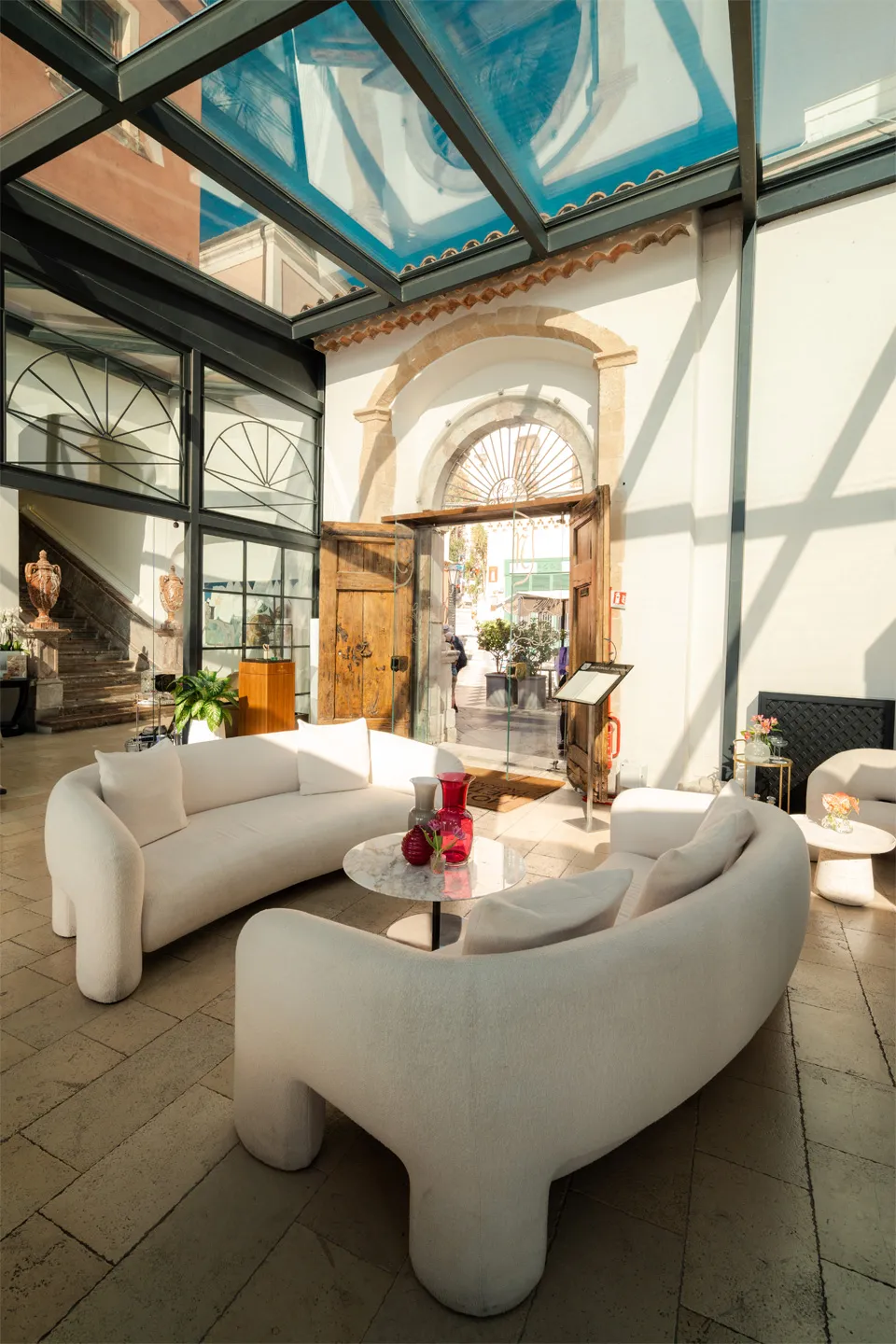 Reception Hotel Metropole Taormina: lobby con divani bianchi, soffitto in vetro, portone in legno antico e arredi di design