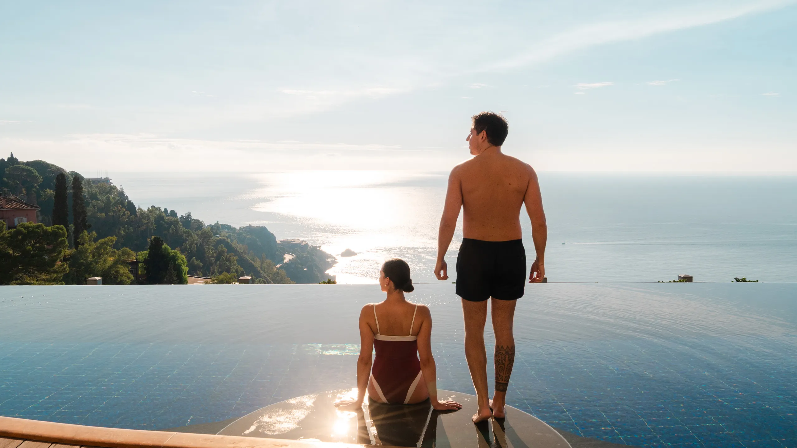 Coppia rilassata bordo della piscina a sfioro dell'Hotel Metropole di Taormina, con vista panoramica sul mare Ionio, la costa siciliana e la vegetazione lussureggiante al tramonto